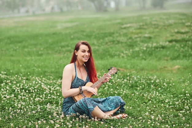 Cheerful woman with red hair playing ukulele in the park sitting on the grass on a summer day.