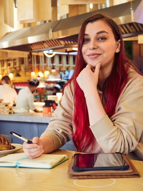 A cheerful woman with red hair works remotely in a cafe writes in a diary