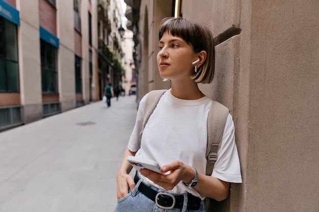 Cheerful woman with smartphone in wireless headphones smiling in city Lady with short hair in white tshirt jeans jeans posing outside on old European street