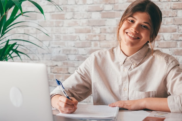 Cheerful woman write notes watch webinar study work on laptop young student learning computer course listening lecture looking and smiling at the cameraxA