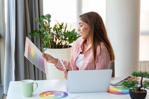 Cheerful youn woman designer having video conference with clients sitting at desk in front of computer holding color palettes gesturing and smiling copy space