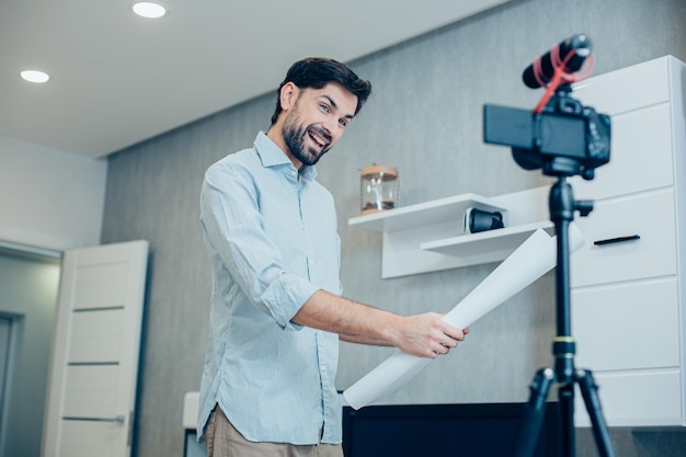 Cheerful young adult man holding a blueprint and smiling to the camera in his living room