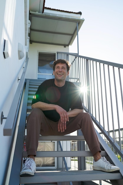 Cheerful young adult man wearing sportswear sitting on the stairs near his house