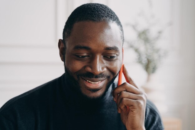 Cheerful young African American businessman talking on smartphone while working distantly at home