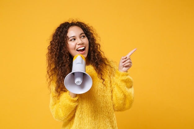 Cheerful young african american girl in fur sweater posing isolated on yellow orange wall background. People lifestyle concept. Mock up copy space. Scream in megaphone, pointing index finger aside.