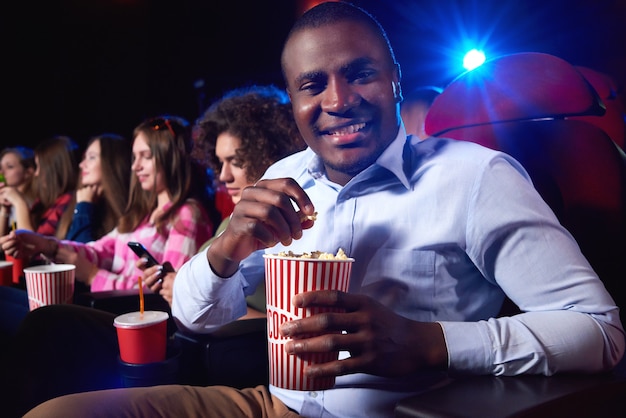 Cheerful young African man smiling holding his popcorn bucket enjoying a movie at the local cinema copyspace snacks happiness entertainment positivity leisure activity concept.