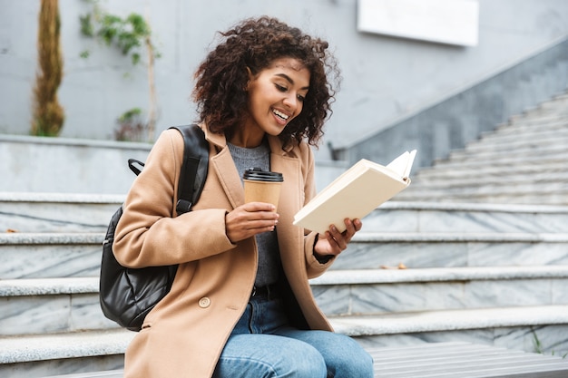 Cheerful young african woman wearing coat sitting outdoors, holding takeaway coffee cup, reading a book