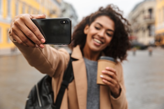 Photo cheerful young african woman wearing coat walking outdoors, holding takeaway coffee cup, taking a selfie