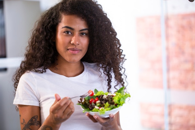 cheerful young afro american woman eating vegetable salad in home kitchen  