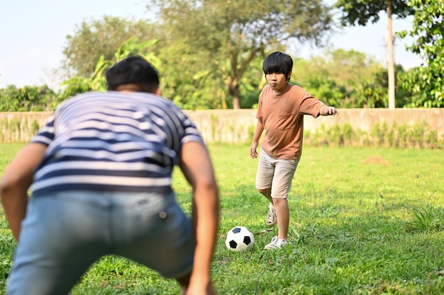 Cheerful young Asian boy playing football with his dad at the backyard having fun time together