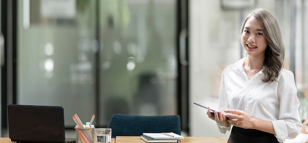 Cheerful young asian businesswoman holding digital tablet smiling and looking at camera standing in modern office