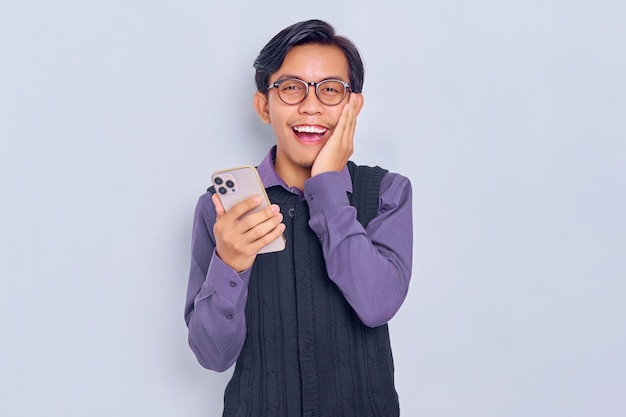 Cheerful young Asian man in casual shirt holding mobile phone and touching her cheek looking at camera isolated on white background