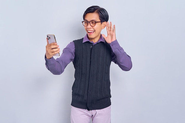 Cheerful young Asian man in casual shirt and vest saying hi and waving hand while making video call on mobile phone isolated on white background People lifestyle concept