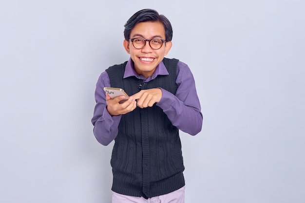 Cheerful young Asian man in casual shirt and vest using smartphone and looking at camera isolated on white background People lifestyle concept