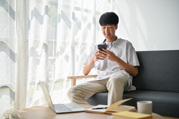 A cheerful young Asian man is scrolling on social media using his smartphone on a sofa