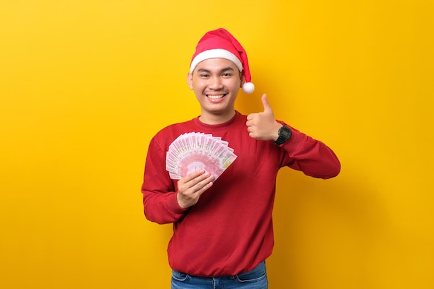 Cheerful young Asian man in Santa hat holding fan of cash money showing thumb up gesture on yellow studio background celebration Christmas holiday and New Year concept