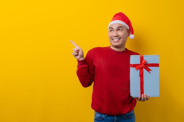 Cheerful young Asian man in Santa hat holding gift box with ribbon pointing finger aside at copy space on yellow studio background celebration Christmas holiday and New Year concept