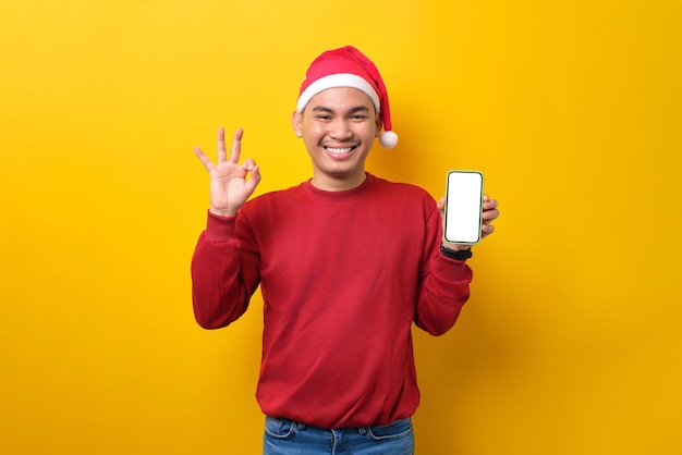 Cheerful young Asian man in Santa hat holding smartphone with blank screen showing okay sign on yellow studio background celebration Christmas holiday and New Year concept