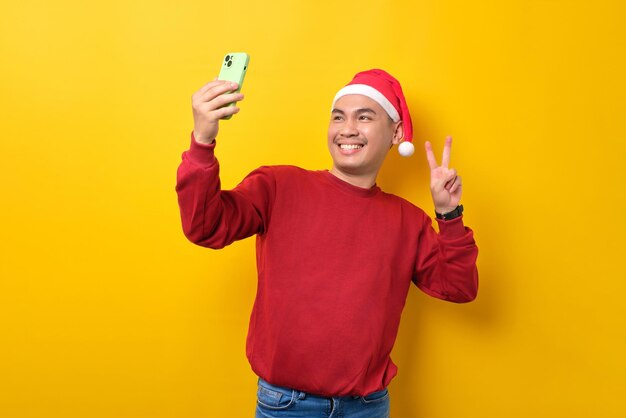 Cheerful young Asian man in Santa hat taking selfie on smartphone showing victory sign over yellow studio background celebration Christmas holiday and New Year concept