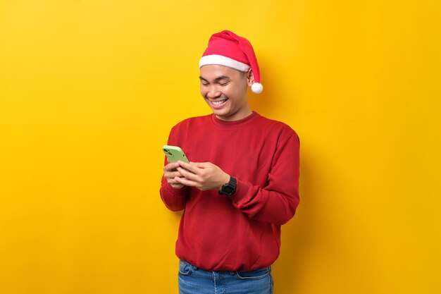 Cheerful young Asian man in Santa hat using smartphone for messaging with friends over yellow studio background celebration Christmas holiday and New Year concept