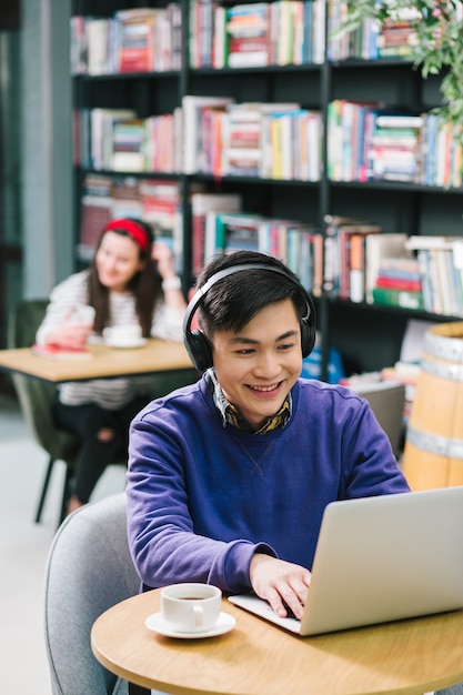 Cheerful young Asian man sitting in a library with headphones on his head and looking at the screen. Cup of coffee on his table