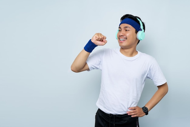 Cheerful young Asian man sporty fitness trainer instructor in blue headband and white tshirt with headphones listen to music sing song in microphone isolated over white background