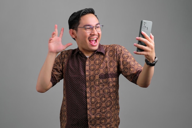 Cheerful young Asian man wearing batik shirt looking at smartphone with excitement and raising hand isolated on grey background