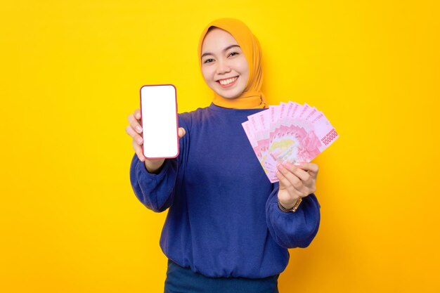 Cheerful young Asian Muslim woman dressed in casual sweater showing blank screen mobile phone and holding money banknotes isolated over yellow background