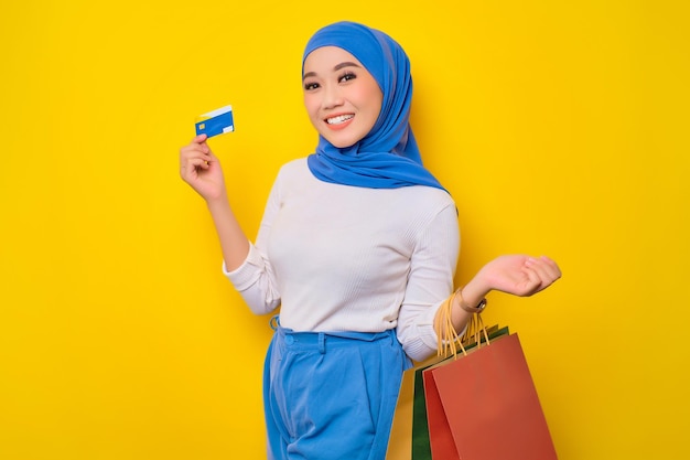 Cheerful young Asian Muslim woman holding credit card and shopping bags looking at camera isolated on yellow background