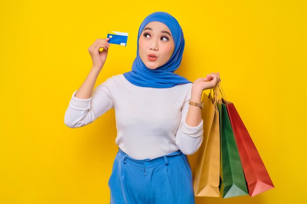 Cheerful young Asian Muslim woman holding credit card and shopping bags looking at camera isolated on yellow background