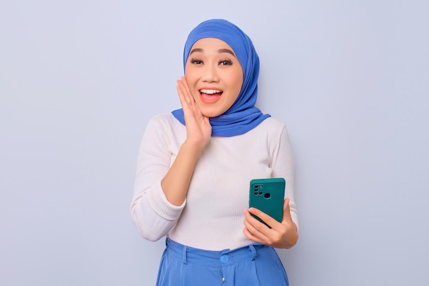 Cheerful young Asian Muslim woman holding mobile phone and looking at camera isolated over white background