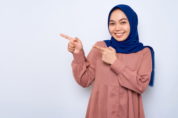 Cheerful young Asian muslim woman in pink shirt pointing fingers at copy space isolated over white background People religious lifestyle concept