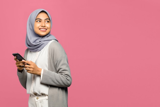 Cheerful young Asian woman holding mobile phone and smiling looking aside