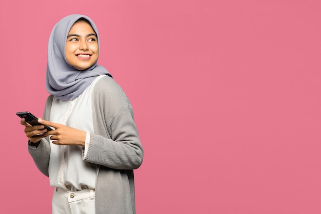 Cheerful young Asian woman holding mobile phone and smiling looking aside