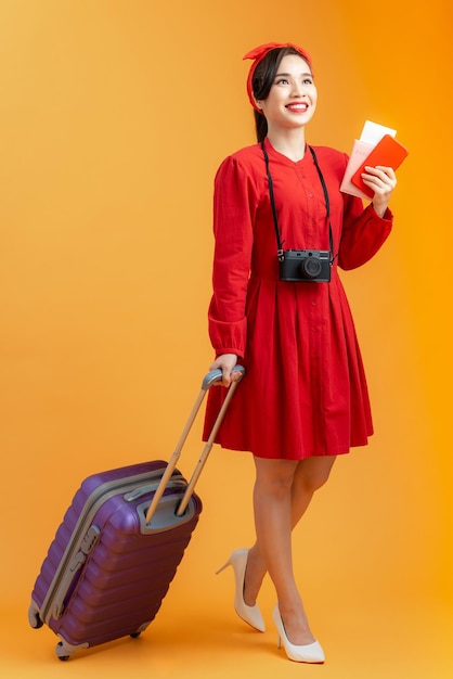 Cheerful young Asian woman holding suitcase passport and flight ticket while standing isolated over orange background