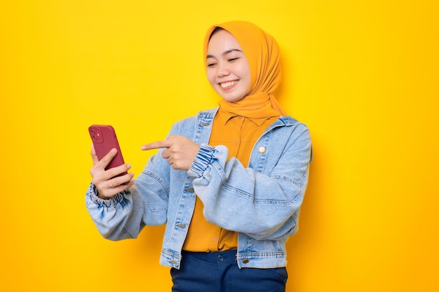 Cheerful young Asian woman in jeans jacket looking at mobile phone screen raised hand with opening mouth reacting to great news isolated over yellow background