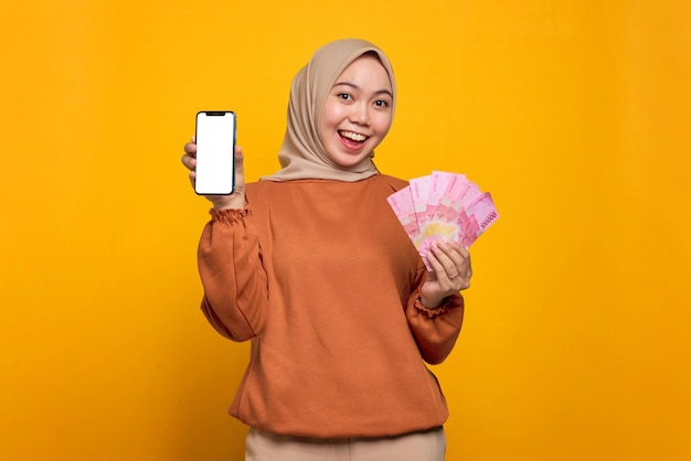 Cheerful young Asian woman in orange shirt showing blank screen mobile phone and holding money banknotes isolated over yellow background