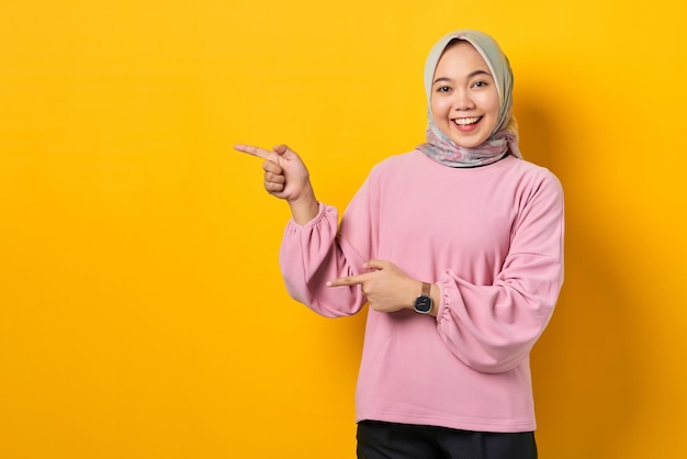 Cheerful young Asian woman in pink shirt pointing fingers at copy space on yellow background