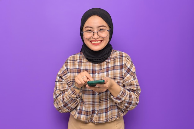 Cheerful young Asian woman in plaid shirt holding a mobile phone getting great news looking at camera isolated on purple background