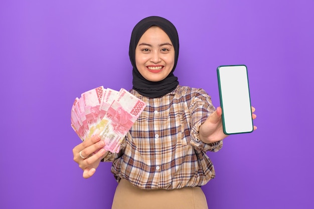 Cheerful young Asian woman in plaid shirt showing blank screen mobile phone and holding money banknotes isolated on purple background