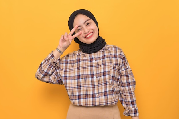 Cheerful young Asian woman in plaid shirt showing peace gesture isolated on yellow background