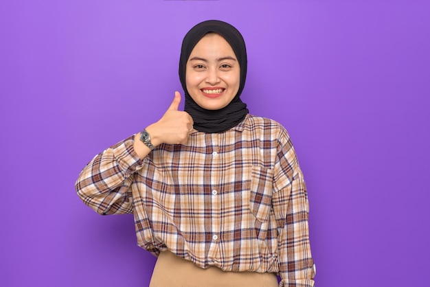 Cheerful young Asian woman in plaid shirt showing thumbs up isolated on purple background