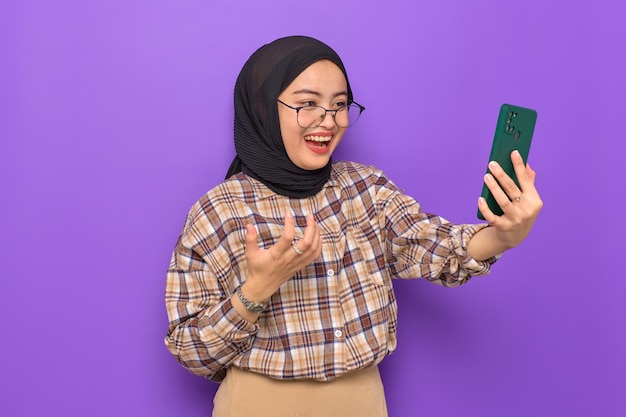 Cheerful young Asian woman in plaid shirt using a mobile phone getting good news isolated on purple background