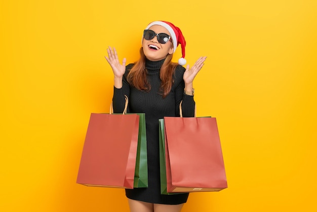 Photo cheerful young asian woman in santa claus hat and sunglasses holding shopping bags and celebrate luck isolated over yellow background