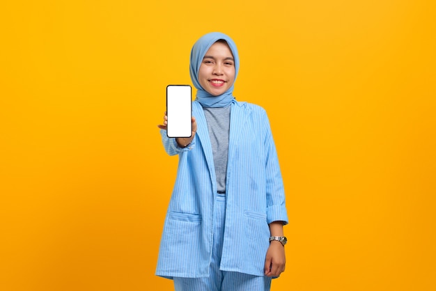 Cheerful young Asian woman showing smartphone blank screen isolated over yellow background