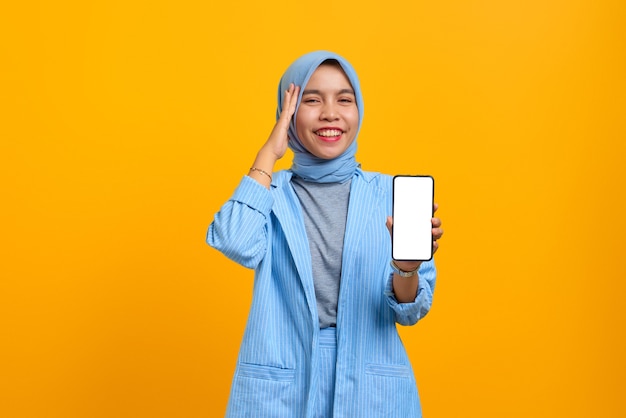 Cheerful young Asian woman showing smartphone blank screen with hand on head over yellow background