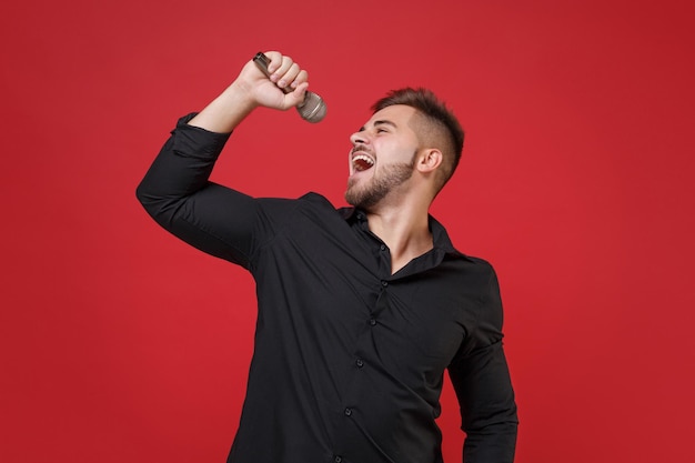 Cheerful young bearded guy in classic black shirt posing isolated on bright red wall background studio portrait. People sincere emotions lifestyle concept. Mock up copy space. Sing song in microphone.