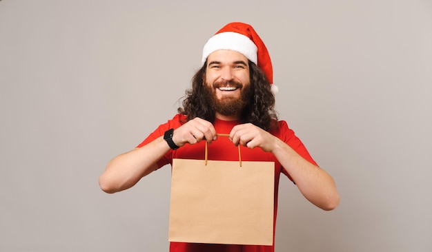 Cheerful young bearded man wearing Christmas hat is holding a paper bag