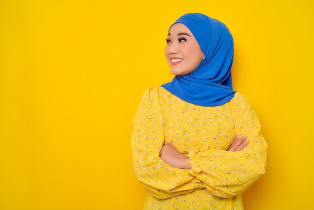 Cheerful young beautiful Asian Muslim woman looking away at copy space with arms crossed isolated over yellow background