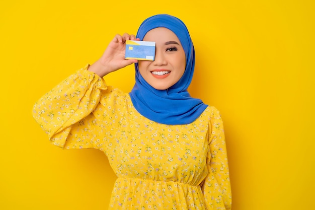 Cheerful young beautiful Asian Muslim woman with Credit Card covering his eye isolated over yellow background